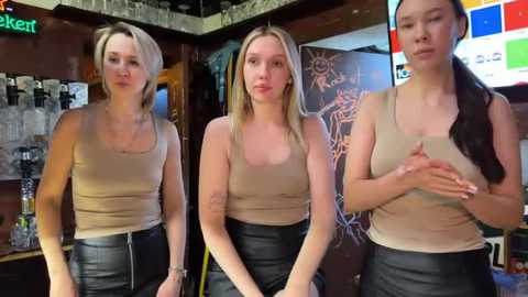 Media: Video of three women in casual beige tops and black skirts, standing in a bar with dark wooden walls, colorful neon lights, and a chalkboard.