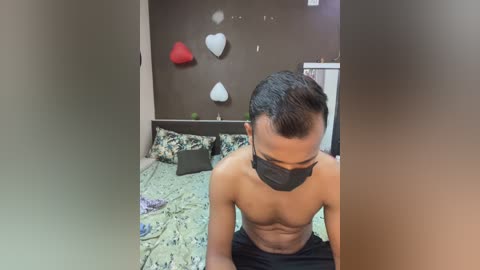 Media: Video of a shirtless man with short black hair and a mask, sitting on a bed with green floral sheets. Background features a dark wall with heart-shaped decorations.