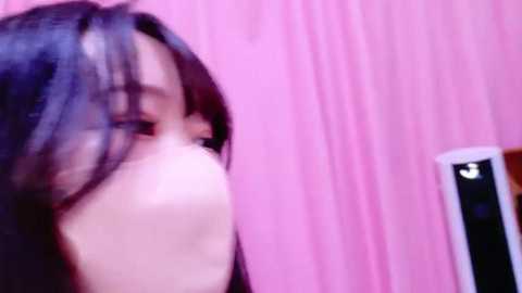 Media: A blurred, close-up video of a young woman with long, dark hair, possibly Asian, against a pink, textured background. She has fair skin and a slender build.