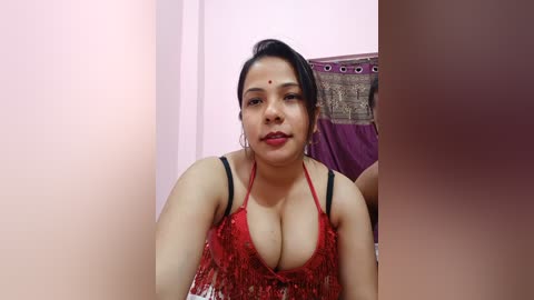 Media: Video of a South Asian woman with fair skin, black hair, and red lipstick, wearing a revealing red lace bra, in a dimly lit room with purple curtains.