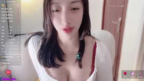 Media: Video of an East Asian woman with fair skin, long black hair, and a nose ring, wearing a white ribbed top, revealing a red bra. Background features a blurred indoor setting with a wooden door.