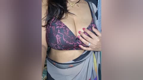 Media: Video of a woman with medium skin tone, wearing a black lace bra with pink floral pattern, revealing ample cleavage. She has long, wavy black hair and is partially covered by a grey and black floral-patterned sarong.