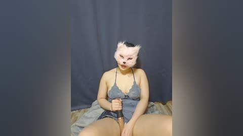 Media: A video of a woman with pale skin, wearing a pink mask and a gray camisole, sitting on a bed with gray sheets, holding a black vibrator between her legs.