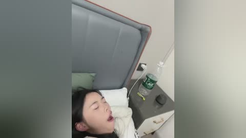 Media: A video shows an Asian woman with dark hair, lying on a hospital bed with a green blanket, mouth open, eyes closed, and a water bottle on a bedside table.