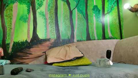 Media: Video of a cozy bedroom with a forest mural on the wall, featuring tall trees, a path, and a beige couch with a green pillow, a beige blanket, and a black bottle on the bed.