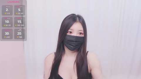 Media: A video of a young Asian woman with long black hair, wearing a black face mask, a black top, and a tattoo on her left shoulder. The background is a plain, white wall.