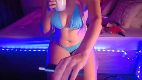 Media: Video of a light-skinned woman with medium breasts in a bright blue bikini, holding a smartphone. Background shows a bed with a stuffed toy and purple LED lighting.