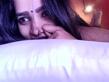 Media: Video of a South Asian woman with long, dark hair, lying on a bed, holding a pillow to her face. The background features a warm, cozy room with a string of fairy lights.