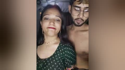 Media: A video of a South Asian woman with long black hair, wearing a green blouse with white dots, smiling, and a shirtless man with glasses in the background, in a dimly lit room.