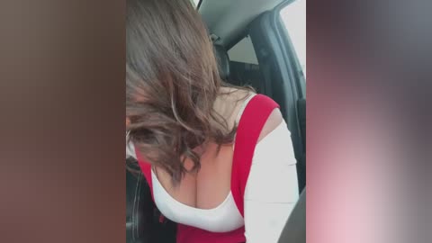 Media: Video of a woman with long brown hair, wearing a red off-the-shoulder top and white long-sleeve shirt, leaning forward in a car. Her large breasts are visible through the low-cut top.