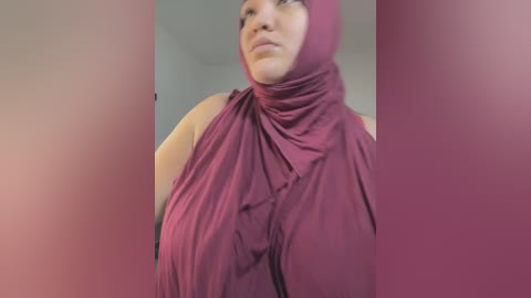 Media: Video of a smiling woman with medium brown skin, wearing a maroon hijab with a large, folded scarf over her head, and a sleeveless top. The background is blurred and gray.