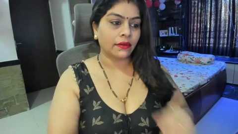 Media: A video of a South Asian woman with medium skin tone, long dark hair, and red lipstick, wearing a black floral-patterned top, sitting on a bed in a dimly-lit bedroom.