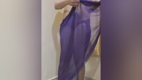 Media: Video of a person holding a sheer, purple cloth in a bathroom with beige walls and a bathtub. The individual's light skin tone is partially visible through the fabric.