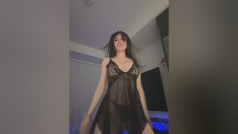 Media: A video of a smiling Asian woman in a sheer black lingerie dress with lace details, standing in a dimly lit bedroom with a flat-screen TV and white walls.