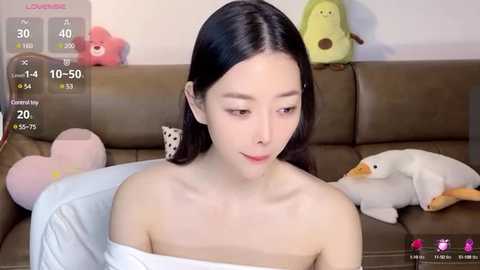 Media: A video of an East Asian woman with long black hair, wearing a white off-shoulder top, sitting on a brown couch. Background includes stuffed toys, a pink bear, and a white duck.