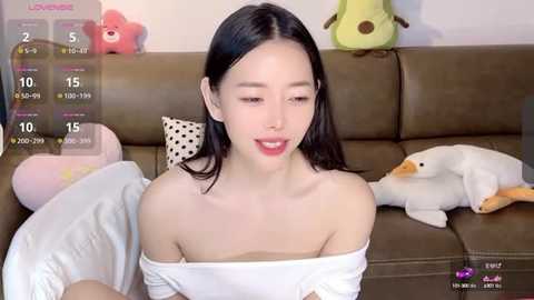 Media: Video of an East Asian woman with fair skin, straight black hair, and a slender physique, wearing a white off-shoulder top. She sits on a brown leather couch, surrounded by plush toys.