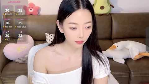 Media: Video of an East Asian woman with long black hair, wearing a white off-shoulder top, sitting on a brown couch. Background includes plush toys and a digital overlay with game statistics.