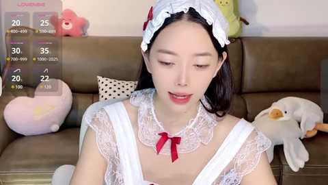 Media: Video of a young East Asian woman with long black hair, wearing a white lace maid outfit with a red bow, sitting on a brown couch with stuffed toys.