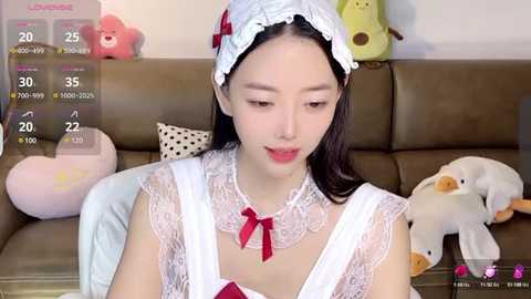 Media: A video of an Asian woman with light skin, wearing a frilly white maid outfit with a red bow, sitting on a beige couch with stuffed toys.