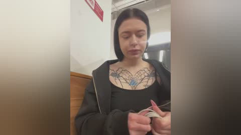 Media: Video of a young woman with fair skin, dark hair, and a black jacket, seated in a subway car. She has a geometric tattoo on her chest, holding a book.