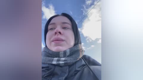 Media: A video of a fair-skinned woman with dark hair, wearing a gray scarf and jacket, taken through a window. She gazes out at a blue sky with scattered clouds.