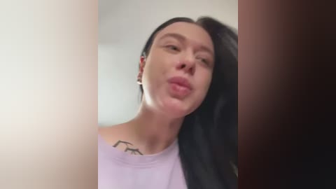 Media: A close-up video of a fair-skinned woman with long black hair, puckering her lips, wearing a white tank top, with a minimalist tattoo on her neck.