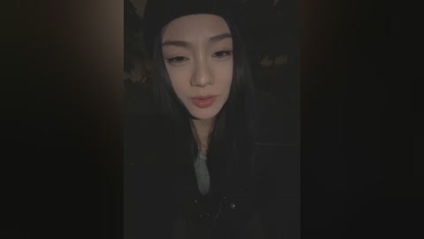 Media: A dimly-lit, close-up video of a young Asian woman with long black hair, fair skin, and red lipstick, wearing a black jacket, taken indoors, likely at night.