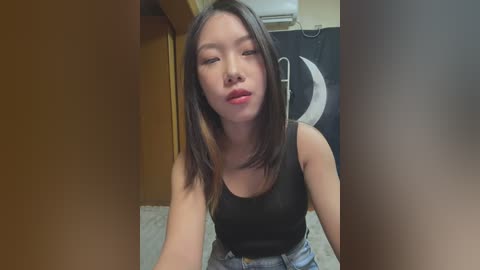 Media: A video of an East Asian woman with long black hair, wearing a black tank top and blue jeans, in a dimly lit room with a black-and-white poster on the wall.