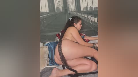 Media: Video of a nude woman with light skin and dark hair, wearing only black lace panties, kneeling on a bed with a beige blanket and red pillow, in a dimly lit room with a poster of the Brooklyn Bridge on the wall.