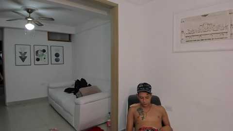 Media: Video of a young, shirtless Black man with tattoos, wearing a headband, sitting on a black couch in a minimalist, white-walled room with abstract art and a ceiling fan.