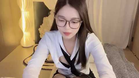 Media: Video of an Asian woman with straight, dark hair, wearing glasses and a white shirt with a black tie. She sits in a cozy room with a striped blanket, lit by a warm, yellow lamp.