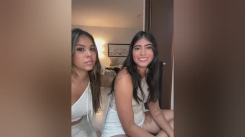 Media: Video of two young women with long, dark hair, smiling, wearing white tank tops and sitting side by side on a bed in a dimly lit room.