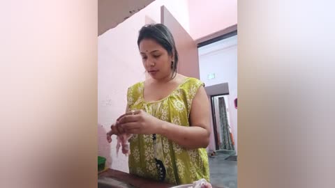 Media: Video of a woman with medium build, medium skin tone, and black hair, wearing a green and white patterned blouse, sitting on a wooden bench, preparing a dish in a simple, pink-painted room with a green curtain.