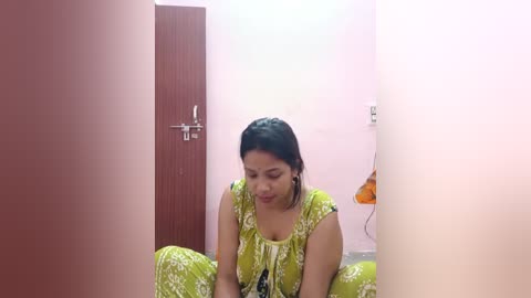 Media: Video of a South Asian woman with medium skin tone, black hair in braids, wearing a green patterned dress, sitting in a pink-walled room with a wooden door and electrical outlets.