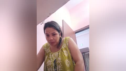 Media: Video of a middle-aged South Asian woman with dark hair and a bindi, wearing a green floral top, standing in a narrow hallway with beige walls and a doorway visible in the background.