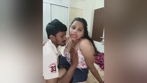 Media: A video shows a young man in a white shirt and blue shorts, with a young girl in a floral top, leaning forward. They are in a modest, beige-tiled room with a bed and a closet.