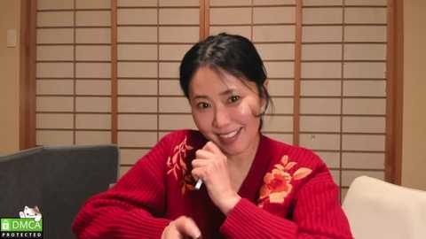 Media: Video of a cheerful Asian woman with dark hair, wearing a red sweater, holding a microphone, in a cozy living room with wooden shutters.