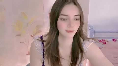 Media: Video of a young Caucasian woman with fair skin, straight brown hair, and light makeup, wearing a floral-patterned top, sitting on a bed with pink sheets and a white headboard.