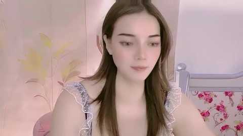 Media: A video of a young Caucasian woman with straight brown hair, fair skin, and a petite build, wearing a white lace top, sitting in a pastel-colored room with floral wallpaper and a pink floral bedspread.