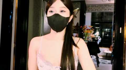 Media: Video of a young East Asian woman with long black hair in a ponytail, wearing a black face mask and a white lace camisole, standing in a modern, well-lit room with a floral arrangement in the background.