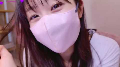 Media: Video of an Asian woman with long, dark hair, wearing a light pink face mask, looking directly at the camera, holding her hair. Background is blurred.