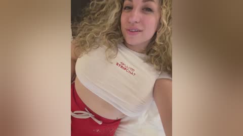 Media: Video of a light-skinned woman with curly blonde hair, wearing a white cropped t-shirt and red shorts, posing indoors with a neutral background.