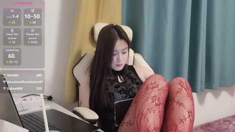 Media: Video of an East Asian woman with long black hair, wearing a black lace top and red lace stockings, seated on a beige gaming chair, typing on a laptop, in a modern, minimally decorated room with teal curtains.