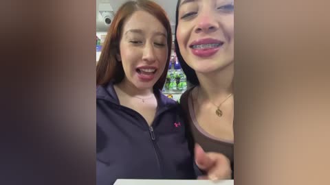 Media: Video of two smiling women in a grocery store. One has fair skin, red hair, and wears a dark jacket; the other has olive skin, black hair, and a beige top. Shelves and products in the background.