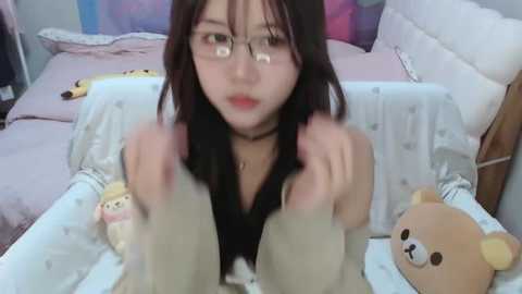 Media: Video of a young Asian woman with glasses and a black choker, in a bed with a stuffed bear, pillows, and colorful wall.