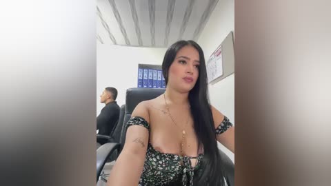 Media: Video of a Latina woman with long black hair, wearing a black floral off-shoulder dress, seated in an office with a male coworker visible in the background.