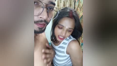 Media: Video of a South Asian man with glasses and a mustache, shirtless, and a woman with red bindi, wearing a striped tank top, in a rustic setting.