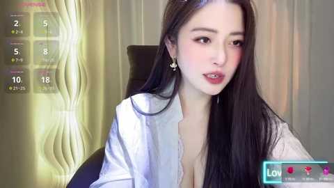 Media: Video of an East Asian woman with long black hair, wearing a white blouse, sitting in a chair. She has fair skin, pink lips, and a neutral expression.