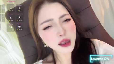 Media: A video of an East Asian woman with long black hair, fair skin, and rosy cheeks, lying on a black chair, with a \"Live on\" overlay and a \"Lovense ON\" watermark.