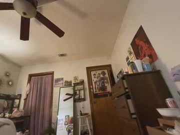 Media: A video of a cluttered bedroom with a ceiling fan, white walls, a TV, wooden furniture, and various personal items.